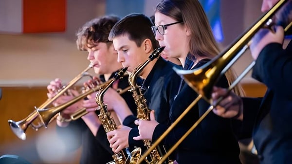 Eine Gruppe Musiker spielt auf der Bühne der Felsted School verschiedene Blech- und Blasinstrumente vor buntem Hintergrund.