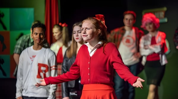 Eine Gruppe Schüler tritt mit roten und weißen Kleidern auf der Bühne vor grünem Hintergrund auf dem Campus des Fingal Community College auf.
