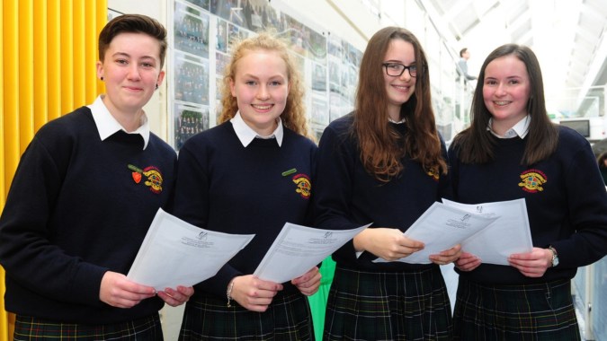 Vier Schülerinnen in Schuluniform stehen vor einer gelben Wand am Fingal Community College und halten Papiere in den Händen.