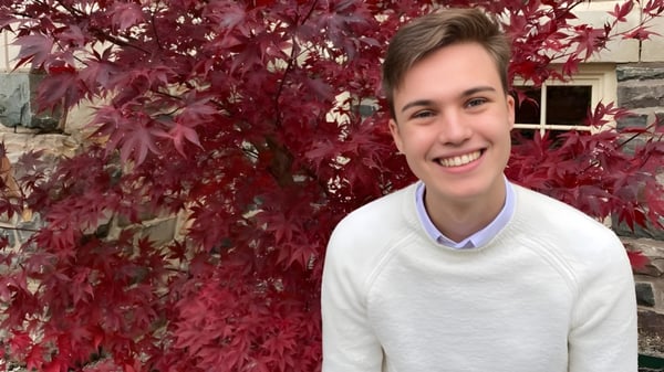 Eine Person mit weißem Pullover steht vor herbstlichem Laub auf dem Gelände der FJ Brennan Catholic High School.