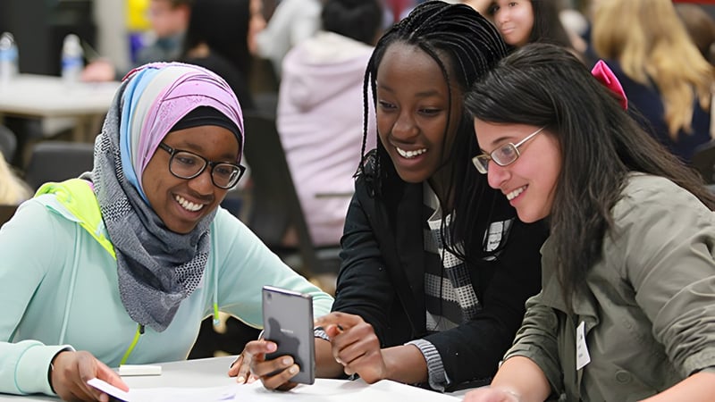 Drei Schülerinnen der Forest Lawn High School schauen gemeinsam auf ein Smartphone im Klassenraum.