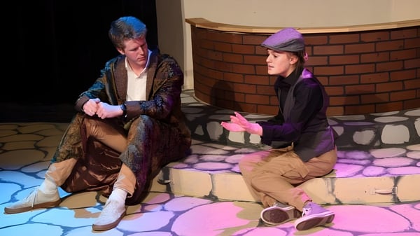 Zwei Personen in farbenfrohen Kostümen sitzen auf der Bühne der Forsyth Country Day School mit Backsteinwand im Hintergrund.