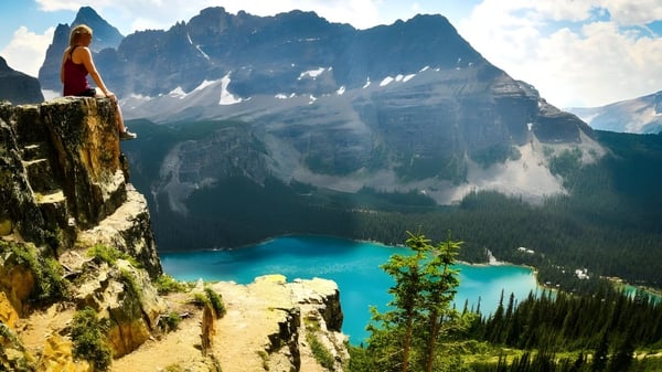 Eine Person blickt von einer felsigen Klippe auf einen türkisfarbenen See und schneebedeckte Berge der Fort Erie International Academy Umgebung.