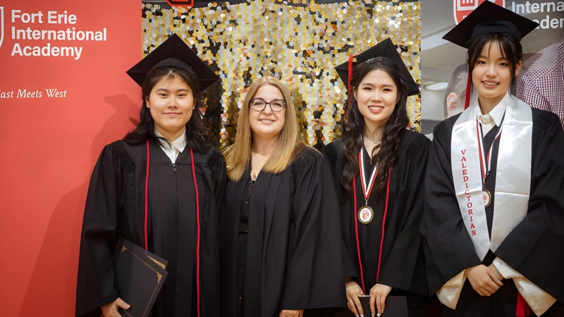 Eine Gruppe von Absolventinnen und Absolventen der Fort Erie International Academy posiert in akademischer Kleidung vor einem Banner mit dem Namen der Schule.