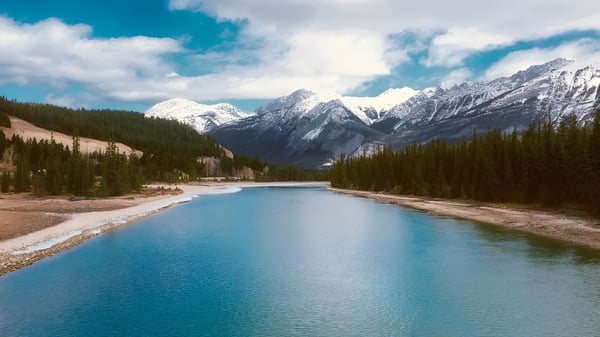 Ein ruhiger türkisfarbener Fluss fließt durch eine Berglandschaft mit schneebedeckten Gipfeln unter bewölktem Himmel.