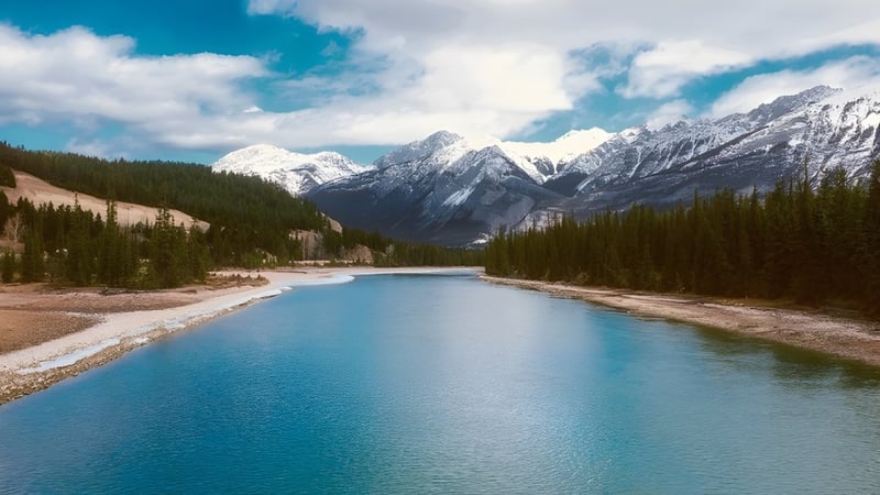 Ein ruhiger türkisfarbener Fluss fließt durch eine Berglandschaft mit schneebedeckten Gipfeln unter bewölktem Himmel.