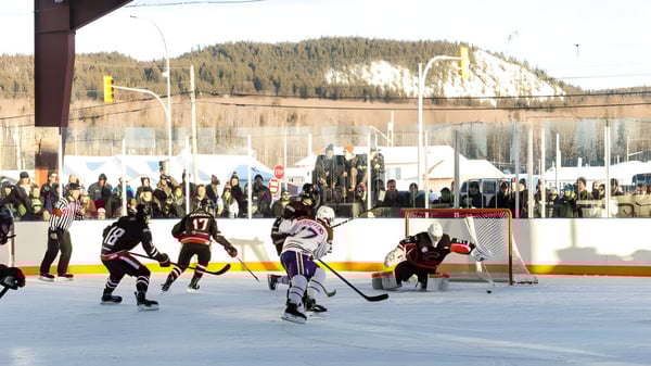 Schüler spielen ein Eishockeyspiel auf dem Außenfeld der Fort St. James Secondary mit Zuschauern und schneebedeckten Bergen im Hintergrund.