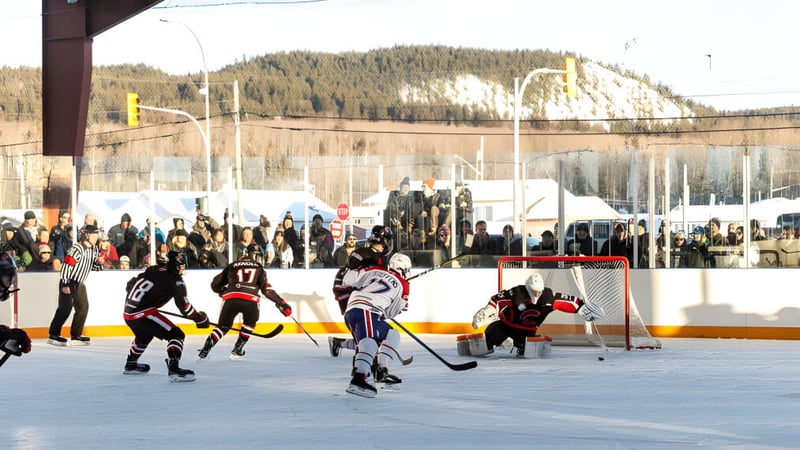 Schüler spielen ein Eishockeyspiel auf dem Außenfeld der Fort St. James Secondary mit Zuschauern und schneebedeckten Bergen im Hintergrund.