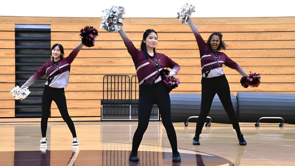 Drei Schülerinnen der Fox Valley Lutheran High School stehen auf dem Basketballfeld in der Sporthalle und üben eine Cheerleading-Routine mit Pompons.