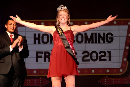 Eine junge Frau mit rotem Kleid und Krone steht mit erhobenen Armen bei der Homecoming-Veranstaltung an der Fox Valley Lutheran High School.