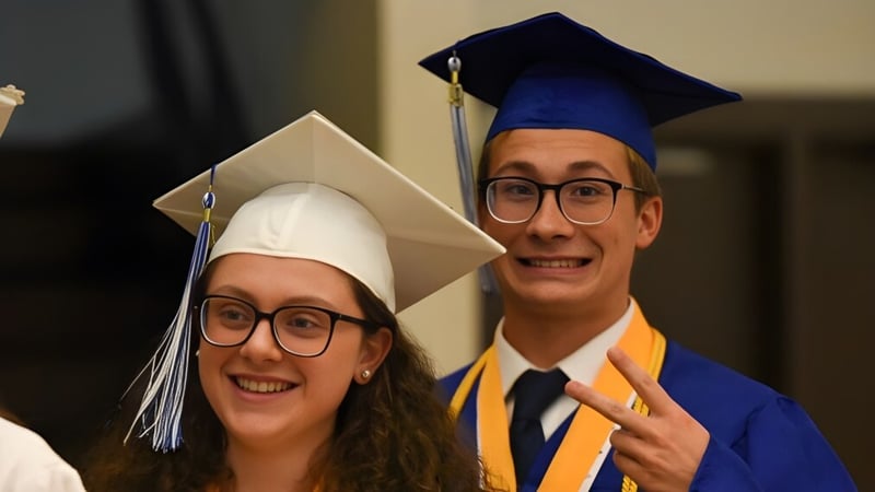 Zwei Absolventen der F.P. Walshe School in Abschlusskleidung stehen zusammen in einem Innenraum.