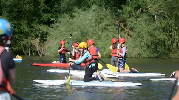 Schüler des Framlingham College paddeln auf Stand-up-Paddleboards auf einem Fluss mit grüner Ufervegetation.
