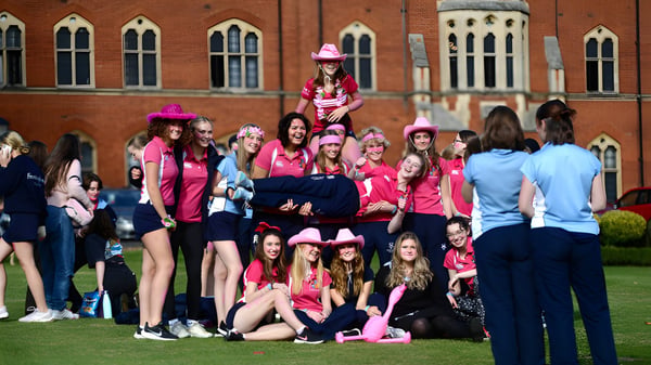 Eine Gruppe Schülerinnen des Framlingham College steht in pinken Outfits auf einer Wiese vor einem ziegelroten Gebäude mit Rundbogenfenstern.