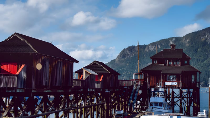 Holzhäuser auf Stelzen über dem Wasser vor Bergen und bewölktem Himmel an der Frances Kelsey Secondary School.