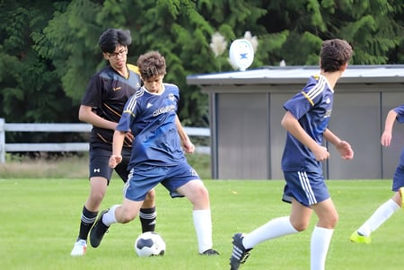 Schüler der Frances Kelsey Secondary School spielen Fußball auf einem grasbewachsenen Feld mit Gebäude und Bäumen im Hintergrund.