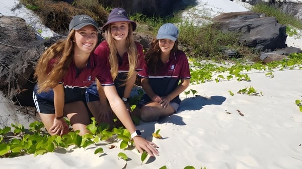 Drei Schülerinnen des Fraser Coast Anglican College stehen am Strand mit Sand, Pflanzen und Felsen im Hintergrund.