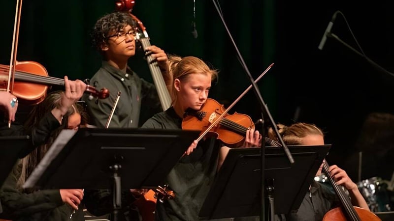 Eine Gruppe von Musikern der Frederick Gunn School spielt auf der Bühne; im Vordergrund ist ein Violinist und ein Mann mit Mikrofon zu sehen.