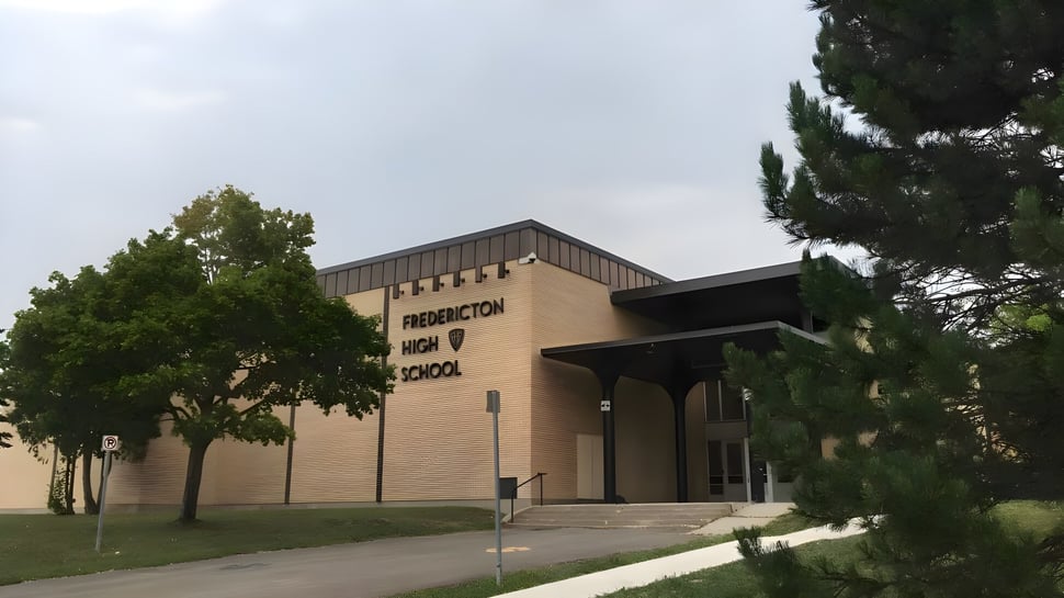 Das Backsteingebäude der Fredericton High School mit Beschriftung am Eingang und einem gepflasterten Weg umgeben von Bäumen.