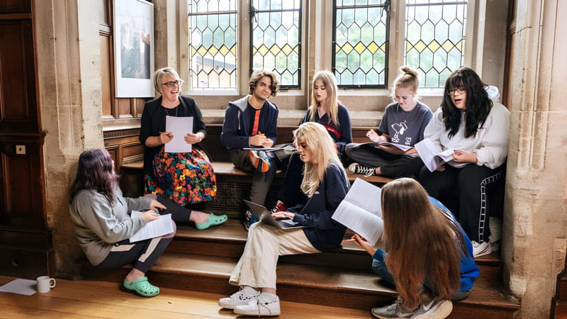Eine Gruppe von Schülerinnen und Schülern sitzt auf den Treppen vor einem alten Gebäude mit bunten Glasfenstern auf dem Campus der Frensham Heights School.