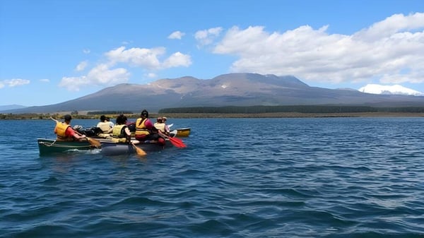 Schüler der Freyberg High School paddeln in einem kleinen Boot auf einem See vor einer Berglandschaft.