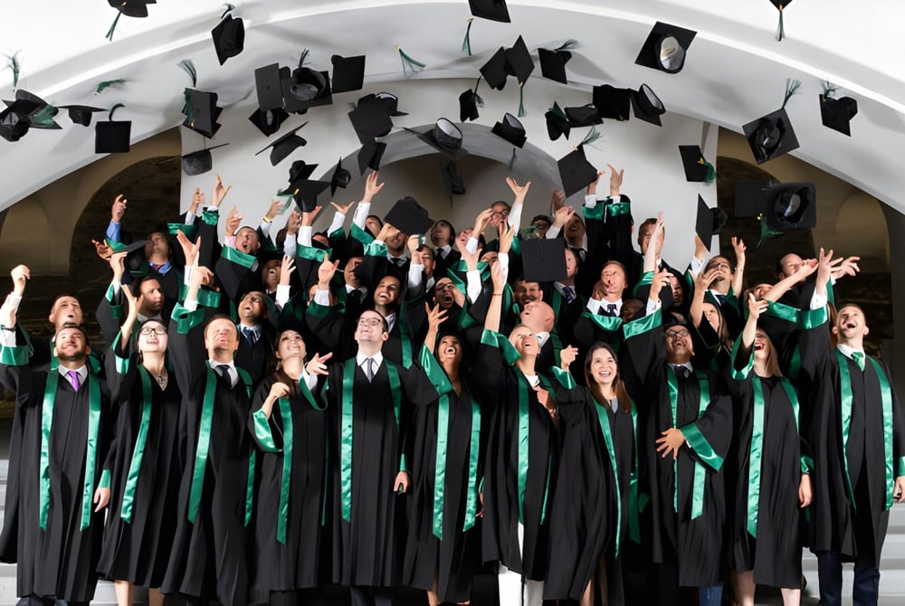 Eine große Gruppe von Absolventinnen und Absolventen der Gallen Community School wirft ihre Abschlussmützen in die Luft vor einem weißen Bogen.