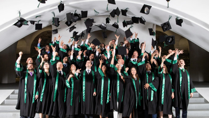 Absolventinnen und Absolventen der Gallen Community School feiern ihren Abschluss mit geworfenen Hüten vor weißem Hintergrund.