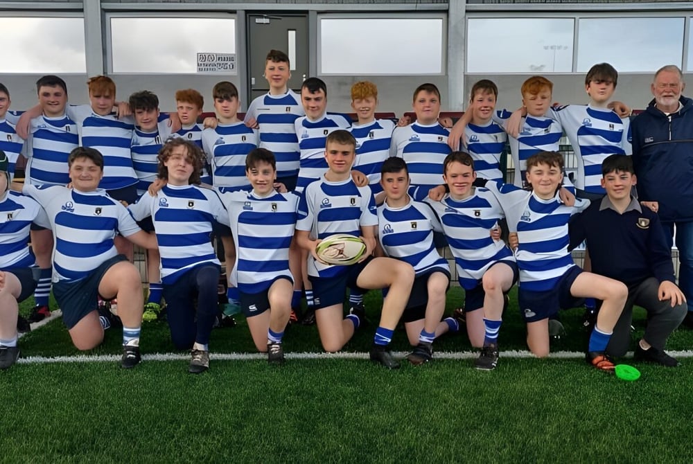 Eine Gruppe junger Rugby-Spielerinnen des Garbally College steht gemeinsam auf einem Spielfeld vor einer Sportanlage.