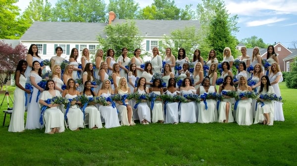 Eine große Gruppe von Frauen in weißen Kleidern mit blauen Akzenten steht auf einer Wiese vor einem Haus auf dem Gelände der Garrison Forest School.