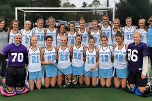 Eine Gruppe Schülerinnen der Garrison Forest School posiert in Sportuniform auf einem Rasenfeld vor Bäumen und Gebäuden.