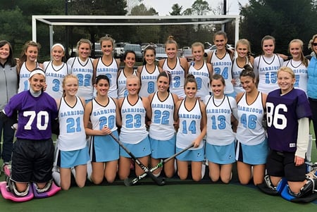 Eine Gruppe Schülerinnen der Garrison Forest School posiert in Sportuniform auf einem Rasenfeld vor Bäumen und Gebäuden.