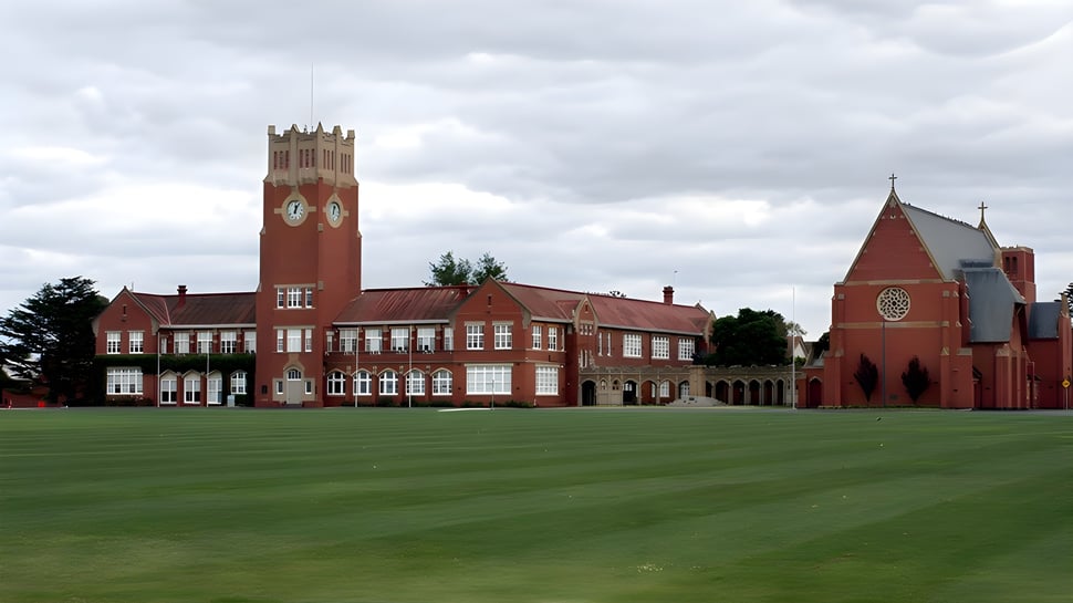Das rote Backsteingebäude mit Uhrturm der Geelong Grammar School steht auf einer grünen Wiese unter bewölktem Himmel.