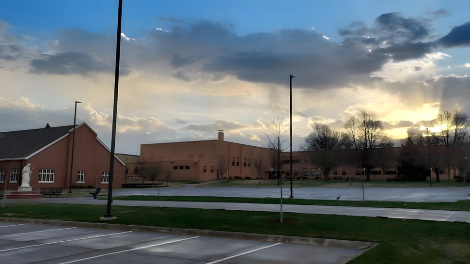 Das Backsteingebäude der Gehlen Catholic School mit einer Wiese im Vordergrund und einem bunten Sonnenuntergang im Hintergrund ist zu sehen.