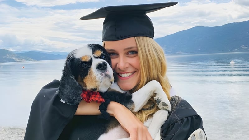 Eine lächelnde Absolventin der George Elliot Secondary School hält einen Hund am Strand mit Bergen im Hintergrund.