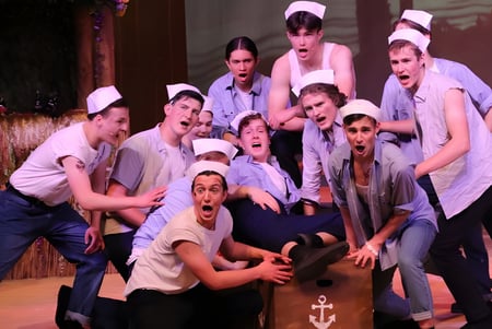 Schülerinnen und Schüler des George Watson’s College in weißen Uniformen bei einer Theateraufführung mit Bambuskulisse.