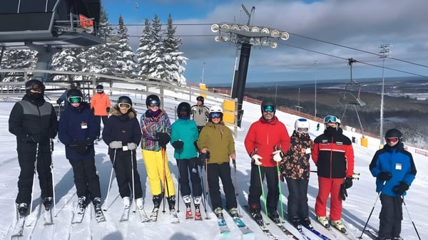 Eine Gruppe von Schülerinnen und Schülern der Georgian Bay District Secondary School steht in bunter Skiausrüstung auf einem schneebedeckten Hang mit Skilift und Bergen im Hintergrund.