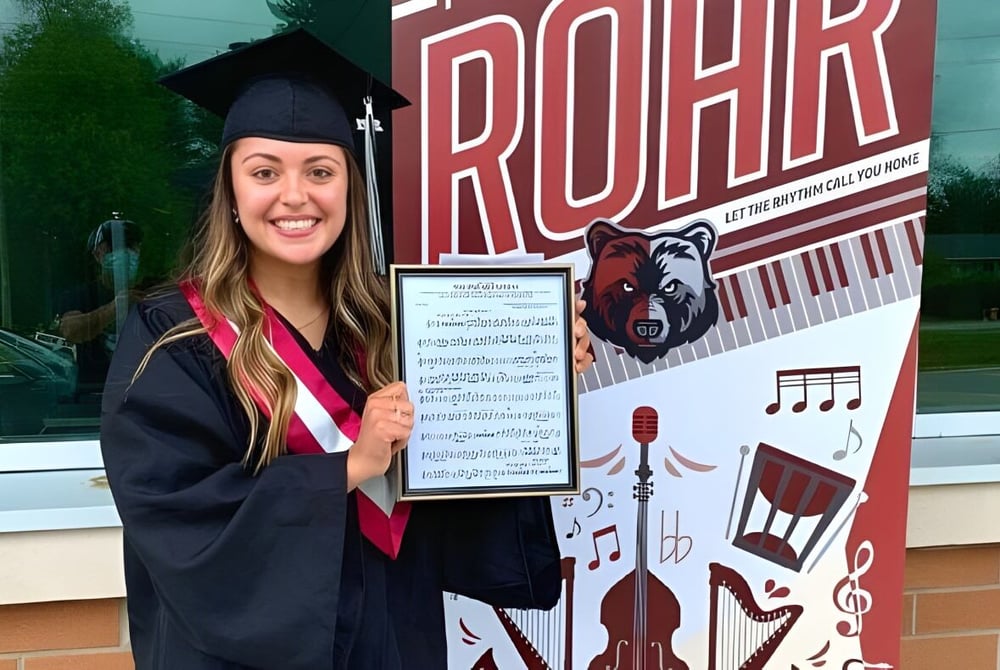 Eine junge Frau in Abschlussrobe steht vor dem großen ROAR-Schriftzug an der Georgian Bay District Secondary School.
