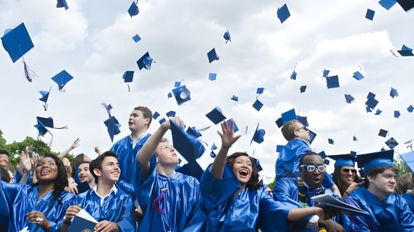 Eine Gruppe von Absolventen der Gilbert Public Schools wirft ihre blaue Abschlussmütze vor wolkigem Himmel in die Luft.