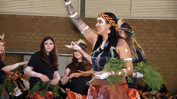 Eine Gruppe von Schülern des Gilmore College führt in bunten, tribal-inspirierten Kostümen auf einer Bühne vor einer Backsteinwand auf.