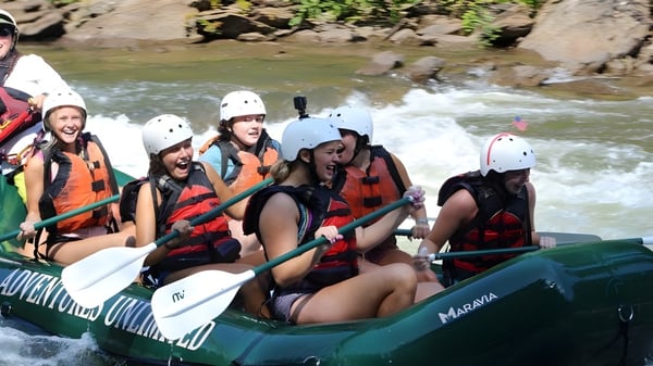 Schülerinnen der Girls Preparatory School sitzen mit Schwimmwesten und Helmen in einem grünen Schlauchboot auf einem Fluss mit felsigem Ufer und dichter Vegetation.