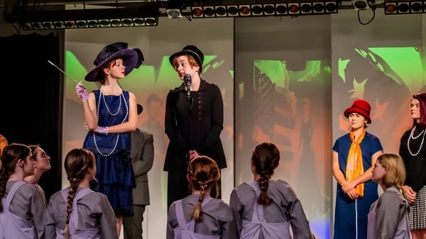 Schülerinnen der Girls Preparatory School führen ein Theaterstück mit historischen Kostümen auf einer Bühne mit bunter Beleuchtung auf.