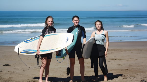 Drei Schülerinnen der Gisborne Girls' High School in Neoprenanzügen mit Surfbrettern am Strand vor dem Ozean.