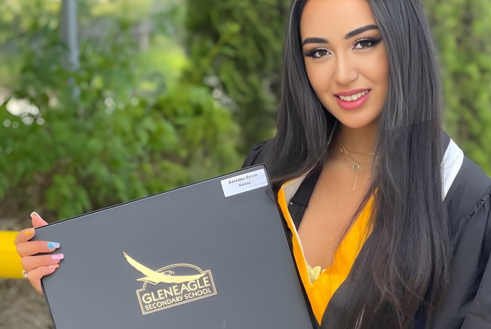 Eine junge Frau hält lächelnd einen grauen Ordner auf dem Campus der Gleneagle Secondary School vor grüner Kulisse.