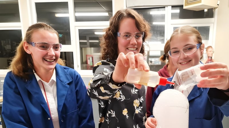 Drei Schülerinnen des Glenfield College stehen lächelnd und gestikulieren in einem Raum mit Fenstern und Regalen.