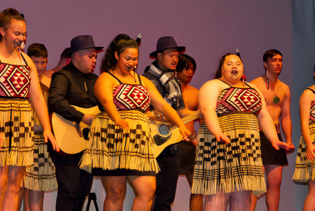 Schüler des Glenfield College führen auf einer Bühne mit lila Hintergrund traditionelle indigene Tänze in traditionellen Kostümen auf.