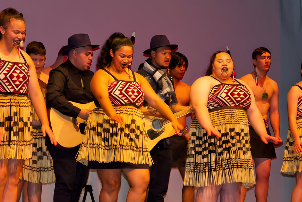 Schüler des Glenfield College führen auf einer Bühne mit lila Hintergrund traditionelle indigene Tänze in traditionellen Kostümen auf.