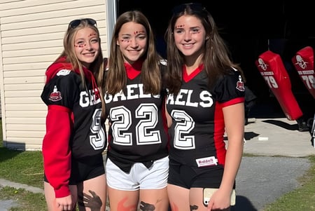 Drei Schülerinnen der Glengarry District High School stehen in einheitlichen Sporttrikots vor einem Holzgebäude.