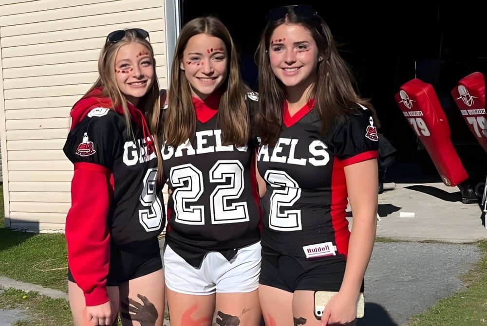 Drei Schülerinnen der Glengarry District High School stehen in einheitlichen Sporttrikots vor einem Holzgebäude.