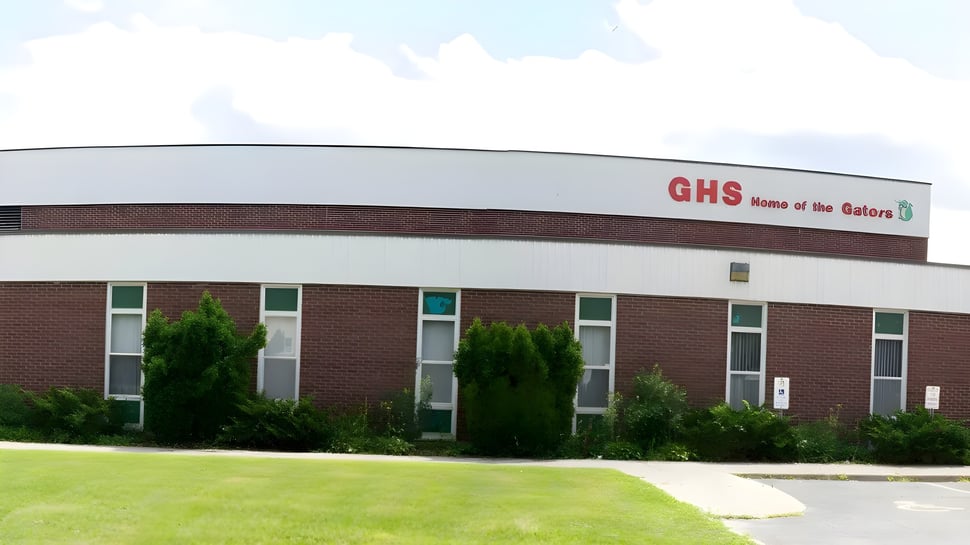 Das große Backsteingebäude der Gloucester High School mit dem Schriftzug Home of the Gators ist von grüner Vegetation und Rasen umgeben.