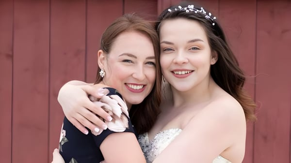 Zwei lächelnde Frauen in formeller Kleidung stehen vor einer roten Holzwand auf dem Gelände der Glovertown Academy.