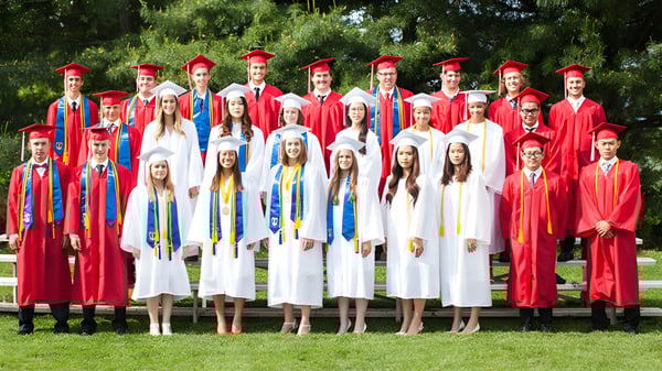 Eine Gruppe von Absolventen der Grace Academy steht in roten und weißen Abschlussgewändern auf einer Wiese vor Bäumen.